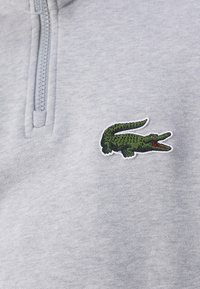 Sudadera gris con cremallera que presenta un logo bordado de cocodrilo verde en el lado izquierdo del pecho. Textura suave y diseño casual.