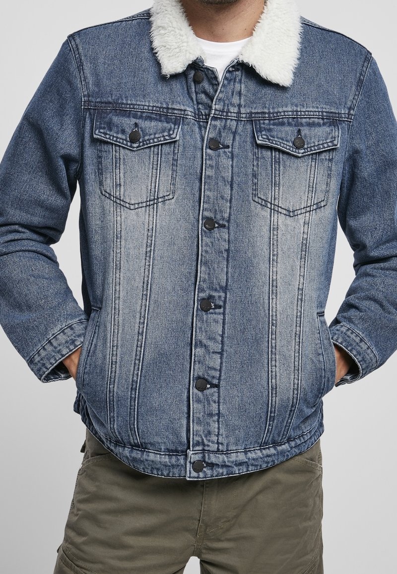 Hombre vestido con una chaqueta de mezclilla azul con cuello de sherpa blanco y pantalones cargo verdes, con las manos en los bolsillos de la chaqueta, de pie contra un fondo liso.