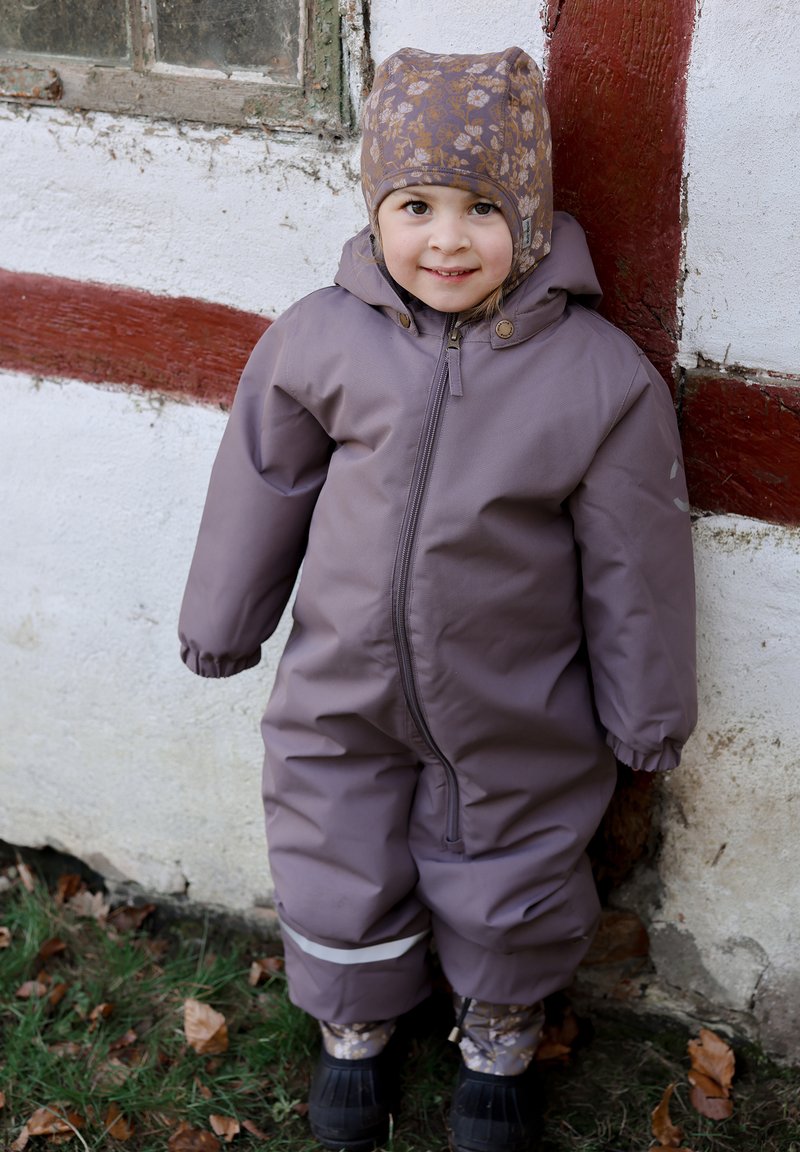 Fialový nepromokavý overal s elastickými manžetami, zipy a reflexními pruhy. Vzorovaná čepice a černé gumové boty doplňují outfit.