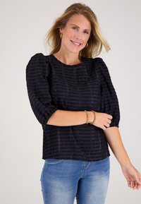 Zwarte blouse met textuur, ballonmouwen en een ronde halslijn, gedragen met lichtblauwe jeans. Gouden armbanden sieren de pols.