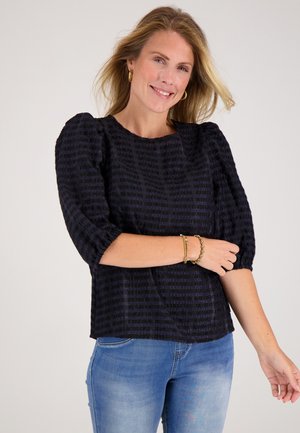 Zwarte blouse met textuur, ballonmouwen en een ronde halslijn, gedragen met lichtblauwe jeans. Gouden armbanden sieren de pols.