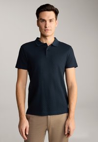 Dunkelmarineblaues kurzärmliges Poloshirt mit strukturiertem Muster, Kragen und Knopfleiste mit drei Knöpfen. Getragen mit hellbeigen Hosen.