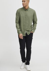 Green button-up shirt with a collar, lightweight fabric, and brown buttons, paired with black pants and white sneakers.