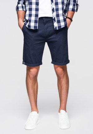 Mężczyzna w granatowych podwiniętych szortach, białych trampkach, białej koszulce i czarno-białej koszuli w kratę, z rękami w kieszeniach.