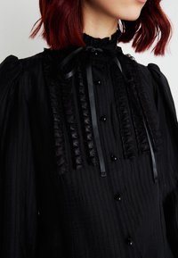 Blouse noire avec un col froncé haut, texture à rayures verticales et accents en ruban satiné. Comprend des boutons noirs et des manches bouffantes.