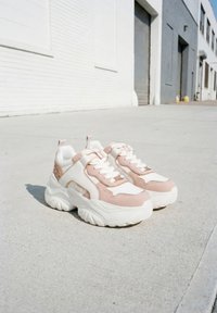 Baskets blanches épaisses avec des touches rose pâle placées sur un trottoir en béton devant des bâtiments industriels blancs et gris.