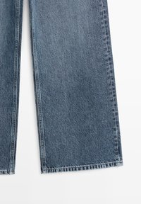 Middenblauwe spijkerbroek met wijde pijpen, zichtbare verticale naden en rechte zomen op een witte achtergrond.