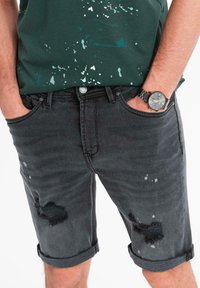 Shorts di denim nero con orli arrotolati, caratterizzati da aree strappate e motivi di vernice schizzata. Indossati con una maglietta verde splatter e un orologio.