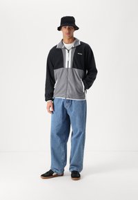 Black and gray zip-up fleece jacket with a high collar, paired with light blue loose jeans and black shoes with brown soles. Bucket hat included.