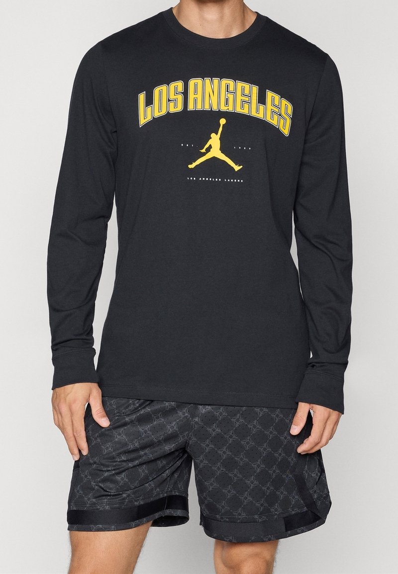 Man wearing black long-sleeve shirt with yellow "Los Angeles" text and basketball player silhouette, paired with black patterned shorts.