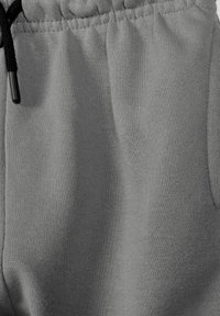 Tissu tricot gris avec ceinture élastique et cordon noir orné d'embouts métalliques, montrant le détail de la poche avant sur un pantalon décontracté.