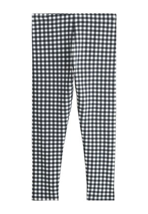 Junges Mädchen, das ein weißes T-Shirt mit Grafik, schwarz-weiß karierte Leggings, weiße Socken und schwarze High-Top-Sneaker trägt, steht vor einem schlichten Hintergrund.
