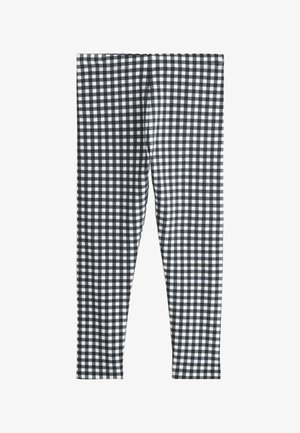 Junges Mädchen, das ein weißes T-Shirt mit Grafik, schwarz-weiß karierte Leggings, weiße Socken und schwarze High-Top-Sneaker trägt, steht vor einem schlichten Hintergrund.