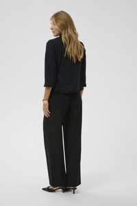 Giacca nera strutturata con orlo arricciato, abbinata a pantaloni neri a gamba larga e décolleté nere con cinturino posteriore. Capelli lunghi e mossi.