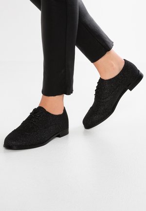 Stopy w czarnych, błyszczących sznurowanych butach, w zestawie z czarnymi, skróconymi spodniami na tle białym.