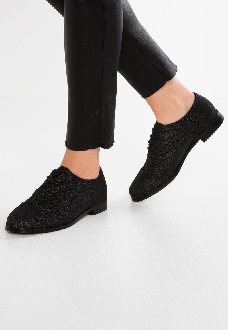 Des pieds portant des chaussures à lacets noires pailletées, associées à un pantalon noir court, sur fond blanc.