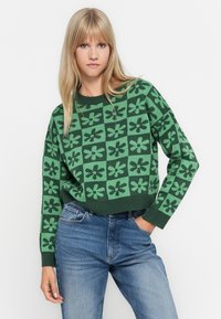 Green patterned knit sweater featuring a floral design in darker green shades. Cropped fit with ribbed cuffs and hem, paired with blue jeans.