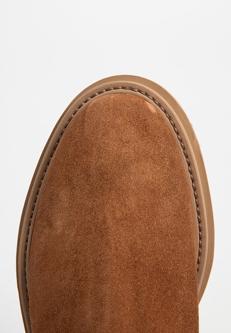 Brauner Wildlederschuh mit detaillierter Naht und tannfarbener Gummisohle am Rand vor einem schlichten weißen Hintergrund.