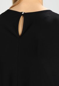 Svart plagg med rund halsringning och en liten nyckelhålssömn på baksidan, med en metallspänne. Slät textur, minimalistisk design.