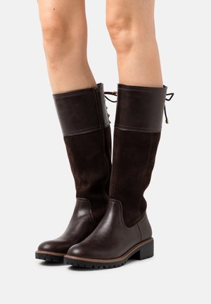 Bottes hautes jusqu'au genou en cuir et suède marron foncé avec fermeture éclair latérale et détail de laçage à l'arrière, portées sur des jambes à peau claire sur fond blanc.