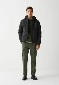 Schwarze Kapuzenjacke mit quilteter Textur, grüner Pullover darunter, olivfarbene Cargo-Hose und schwarze Schuhe. Minimalistisches Design, figurbetonte Silhouette.
