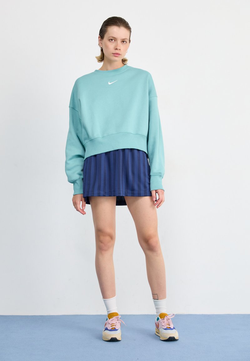 Lichtblauwe cropped sweatshirt met een wit logo, gecombineerd met een navy geplooide rok. De voeten zijn versierd met pastel sneakers en witte crew sokken.