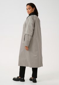 Ljusgrå vattentät trench coat med svart krage, långa ärmar, sidofickor och rak skärning. Modell visad bakifrån.