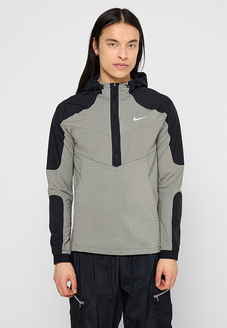 Veste Nike grise avec des accents noirs, dotée d'une fermeture zippée mi-hauteur, d'une capuche, de panneaux texturés et d'un design ajusté. Logo réfléchissant sur la poitrine.
