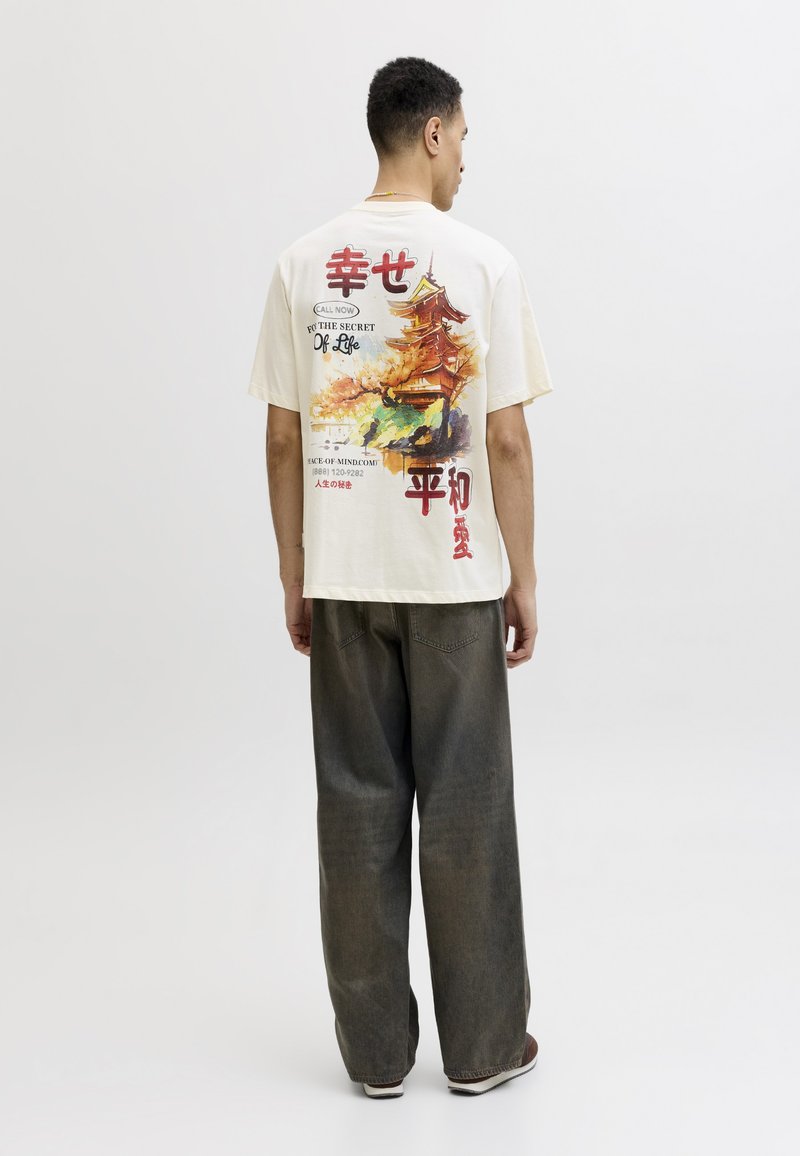 Bărbat stând cu spatele, purtând un tricou alb cu o pagodă și text japonez, asortat cu pantaloni largi închiși la culoare și adidași pe un fundal simplu.
