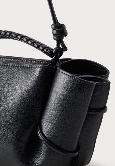 Μαύρη δερμάτινη τσάντα χειρός με πλεκτή λαβή και λεπτομέρεια δεμένου ιμάντα, που εμφανίζει υφή και ραφές σε απλό ανοιχτό φόντο.