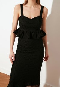 Abito in pizzo nero con silhouette sagomata, vita peplo, scollatura quadrata e spalline larghe. Presenta un tessuto testurizzato con motivi floreali.