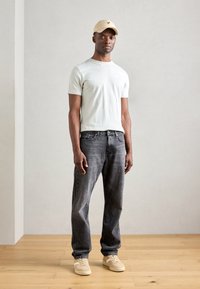Camiseta de manga corta verde claro, jeans negros desgastados y zapatillas beige. Gorra beige con logo. Fondo neutro y suelo de madera.