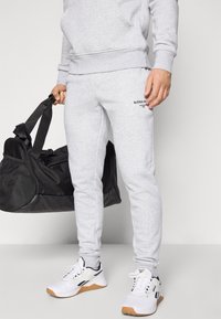 Light grey sweatpants with black logo detail, paired with white athletic shoes and a black duffel bag featuring mesh accents.