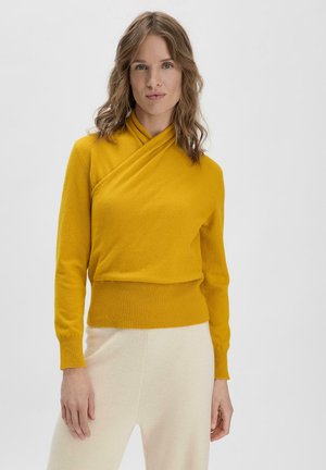 Mosterdgele wikkeltrui met een kruisoversluiting, lange mouwen en een geribde zoom. Gladde textuur, gecombineerd met lichtgekleurde broek.