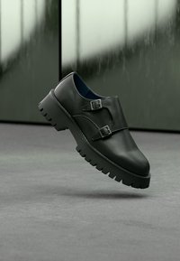 Μαύρα δερμάτινα παπούτσια με στρογγυλεμένη μύτη, δύο λουριά με αγκράφες και χοντρή λαστιχένια σόλα. Λείος υφή με διακριτική λάμψη.