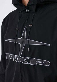 Giacca nera con cappuccio e zip, estremità del cordino argentate e grande logo stilizzato a forma di stella con testo "PXPP" sul petto.