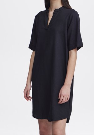 Kobieta w luźnej, czarnej sukience do kolan z krótkimi rękawami i małym kołnierzykiem typu mandarynka z dekoltem w kształcie litery V.