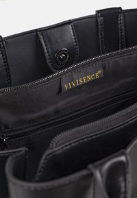 Innenraum einer schwarzen Ledertasche mit Reißverschlusstasche, Druckknopfklappe und Stoffetikett mit goldener "VIVISENCE"-Markenkennung.