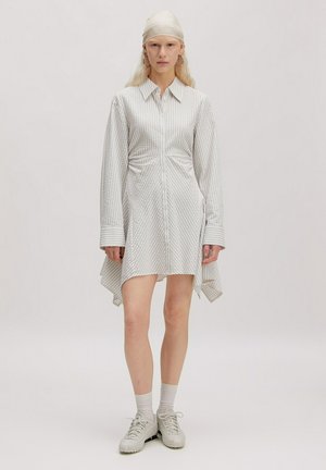 Femme portant une robe-chemise blanche à manches longues avec des rayures diagonales, des baskets blanches, des chaussettes, des boucles d'oreilles créoles et un foulard blanc, debout devant un fond uni.