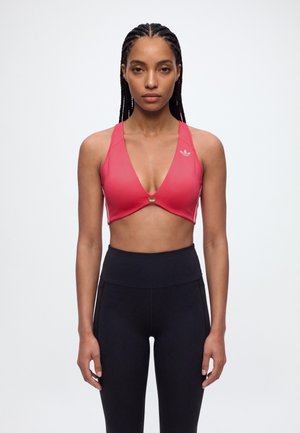 Une femme aux cheveux tressés se tient face à l’objectif, vêtue d’un soutien-gorge de sport rose et d’un legging noir, sur un fond clair et uni.