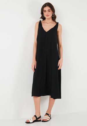 Black sleeveless dress with a V-neck, knee-length, and soft fabric. Notable knot accents on shoulders; paired with black and white sandals.