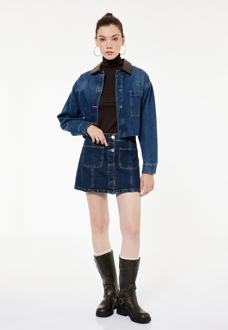 Veste en denim avec col marron, manches courtes et poche avant, associée à une mini jupe à boutons. Le mannequin porte des bottes marron dépassant le genou.