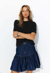 Pull noir à manches courtes avec une finition texturée, associé à une jupe en denim bleu moyen avec un ourlet à volants et des coutures visibles.