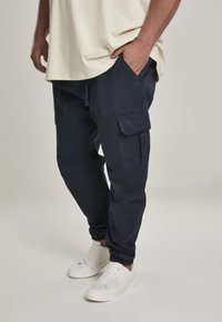 Navy cargo pants with elastic cuffs, drawstring waist, and multiple pockets, paired with white sneakers and a loose beige t-shirt.