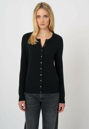 Frau trägt eine schwarze, mit Knöpfen versehene Strickjacke und dunkelgraue Jeans, stehend vor einem einfarbigen weißen Hintergrund.