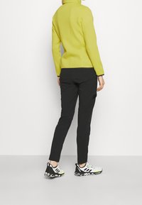 Gelbe Fleecejacke mit hohem Kragen, schwarze Cargo-Hosen und schwarz-weiße sportschuhe mit neongelben Akzenten. Rückansicht gezeigt.