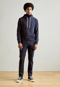 Sudadera azul marino con cordones blancos y bolsillo frontal, combinada con pantalones cargo azul marino oscuros y zapatillas negras con suelas blancas.