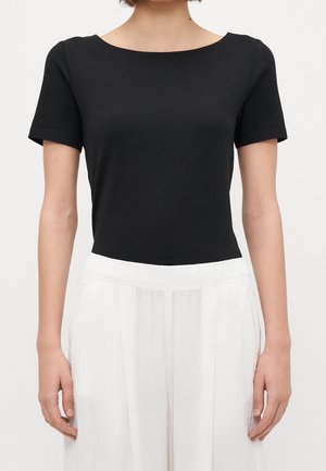 Frau trägt ein schlichtes schwarzes Kurzarmshirt, das in lockere, hoch taillierte weiße Hosen gesteckt ist, vor neutralem Hintergrund.