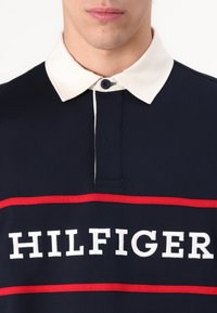 Navy cotton polo shirt with a cream collar, featuring embroidered "HILFIGER" text and a red stripe across the chest.