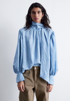 Hellbluse mit einer knitterigen Textur, hohem Ausschnitt und langen Puffärmeln. Kombiniert mit lockeren braunen Hosen.